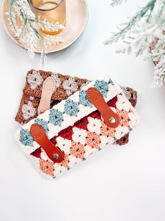 Two crocheted pouches with leather straps are placed on a white surface, accompanied by a glass of liquid and some decorative greenery, making for a charming handmade gift.