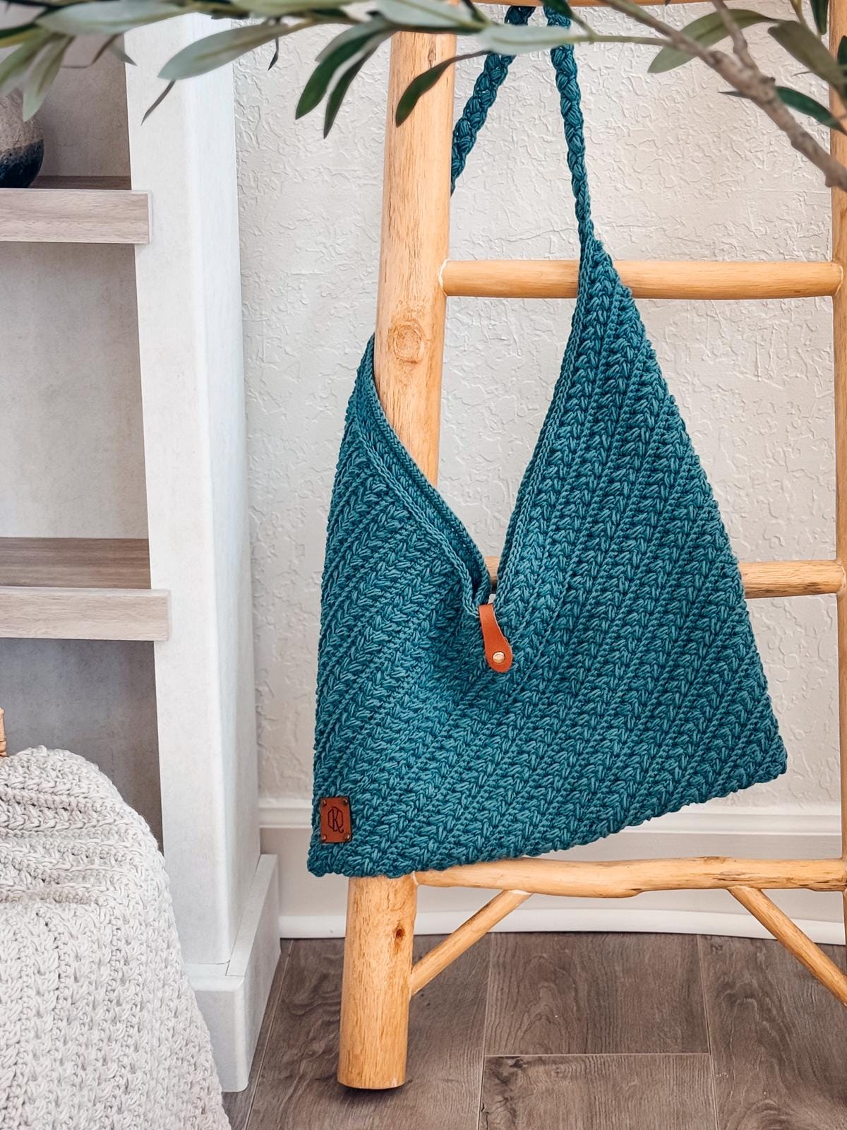 A teal crocheted shoulder bag with a leather tag and button hangs on a wooden ladder near a textured white wall and knit pouf—perfect as a handmade gift or to pair with vintage clothes for added charm.
