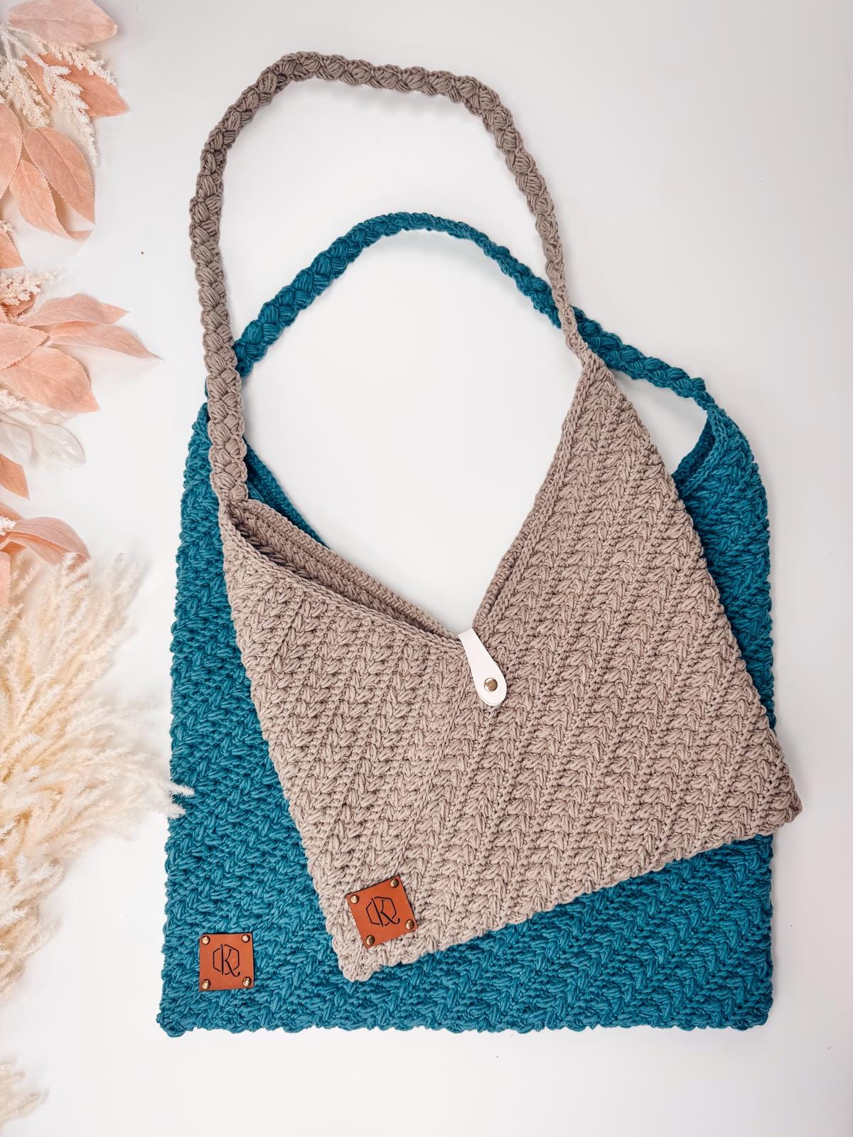 Two crocheted bags, one teal and one beige, each with a textured pattern and a small brown tag, are displayed overlapping on a white surface with decorative foliage nearby—perfect as a handmade gift or to pair with vintage clothes.