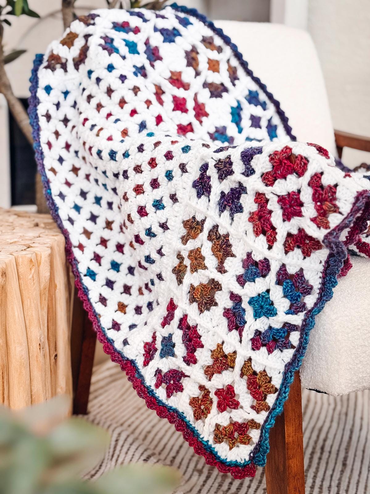 A colorful crocheted granny square blanket with a white base and multicolored squares is draped over a chair next to a wooden stump table, making a perfect handmade gift for lovers of cozy décor.