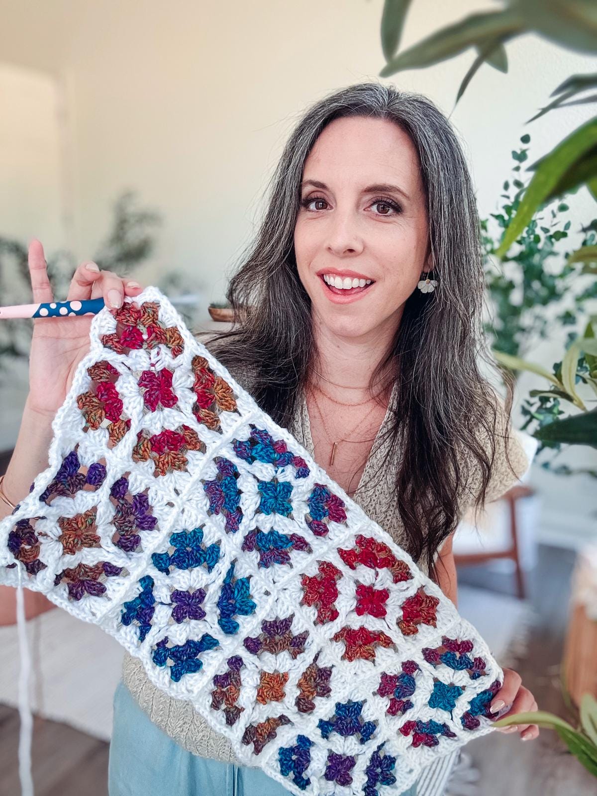 A woman holds up crochet granny squares made of colorful floral patterns with a white border, smiling at the camera in a bright indoor setting—her creation would make a charming handmade gift.