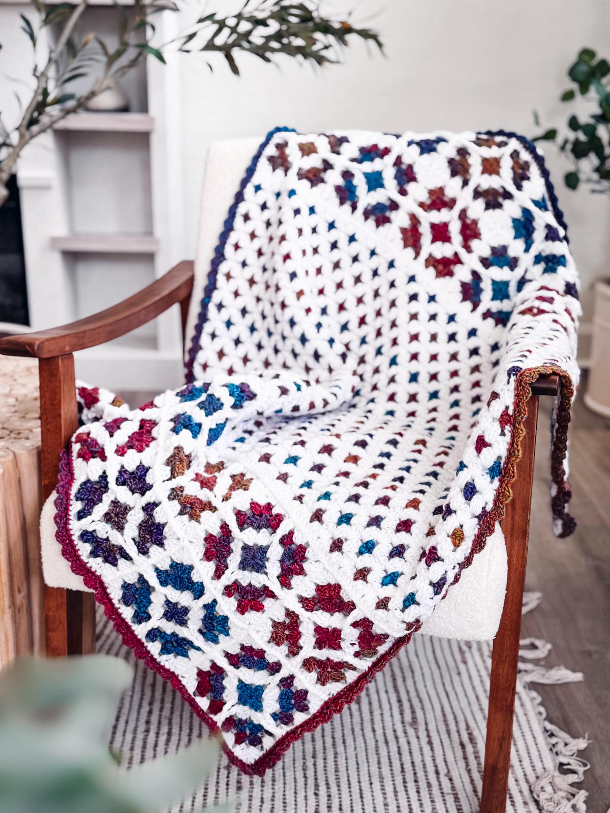 A crocheted blanket with a colorful floral pattern is draped over a white armchair in a bright, cozy room, making the perfect handmade gift for anyone who loves vintage clothes and unique style.