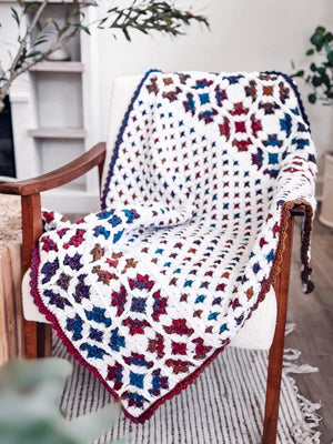 White crochet blanket with jewel-tone granny motifs and a dark border draped over a white armchair