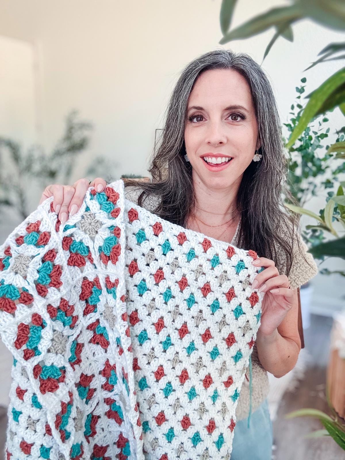 A woman with long brown and gray hair holds up a crocheted blanket with a white, teal, and rust-colored pattern, standing indoors among plants and wearing unique jewellery—a perfect scene for a handmade gift.