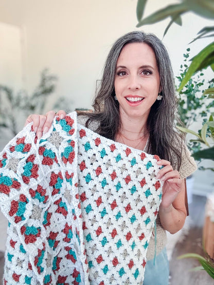 Smiling woman holding up a white crochet blanket with teal, rust, and tan granny motifs among houseplants