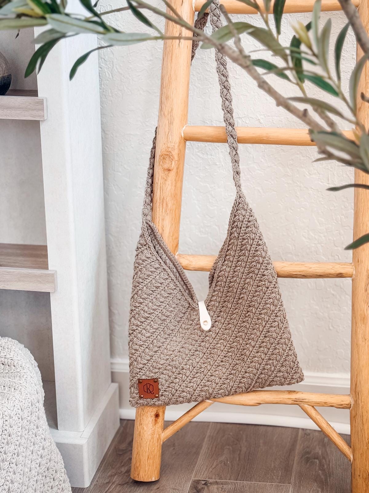 A beige crochet shoulder bag with a button closure hangs on a light wooden ladder, perfect as a handmade gift. Part of a plant is in the foreground and shelves are visible in the background.