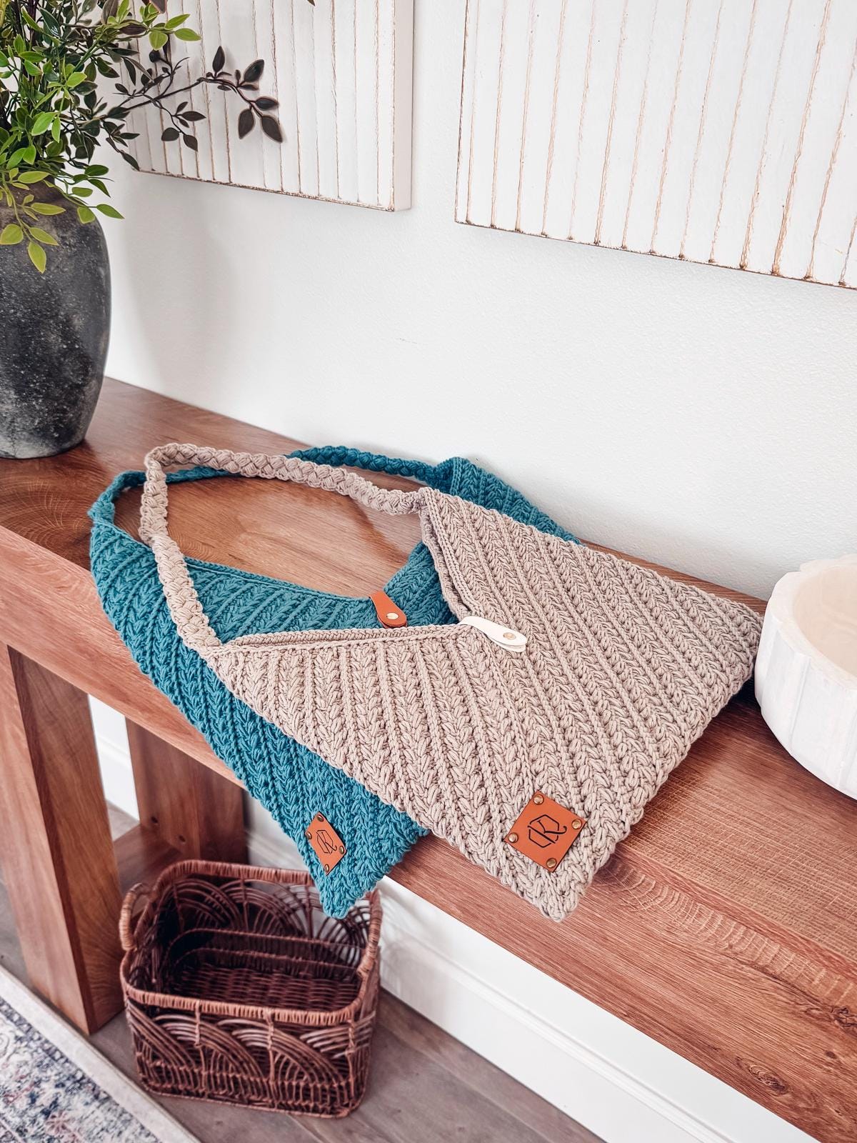 Two crochet tote bags, one beige and one teal, rest on a wooden console table next to a black vase with greenery and a decorative white bowl—ideal as a daily crochet purse.