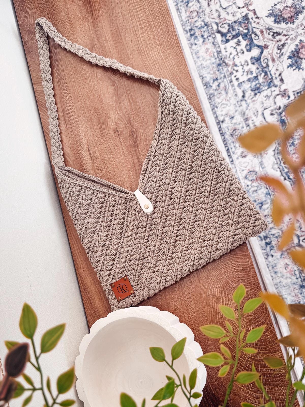 A beige knitted shoulder bag with a button closure sits on a light wooden table, perfect for a crochet project. Part of a plant is in the foreground and shelves are visible in the background.