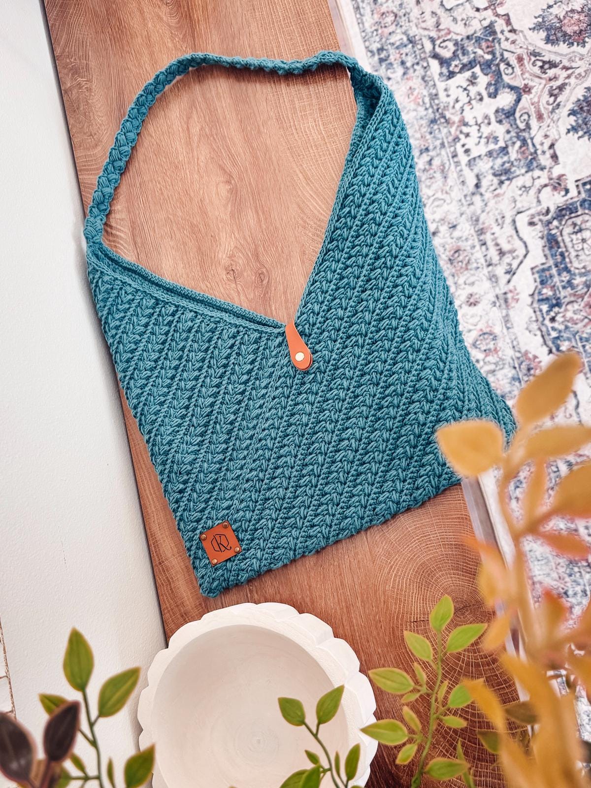 A teal crochet shoulder bag with a textured pattern and leather tag rests on a wooden surface, near a white bowl and leafy plants—an ideal handmade gift for lovers of vintage clothes and unique jewellery.