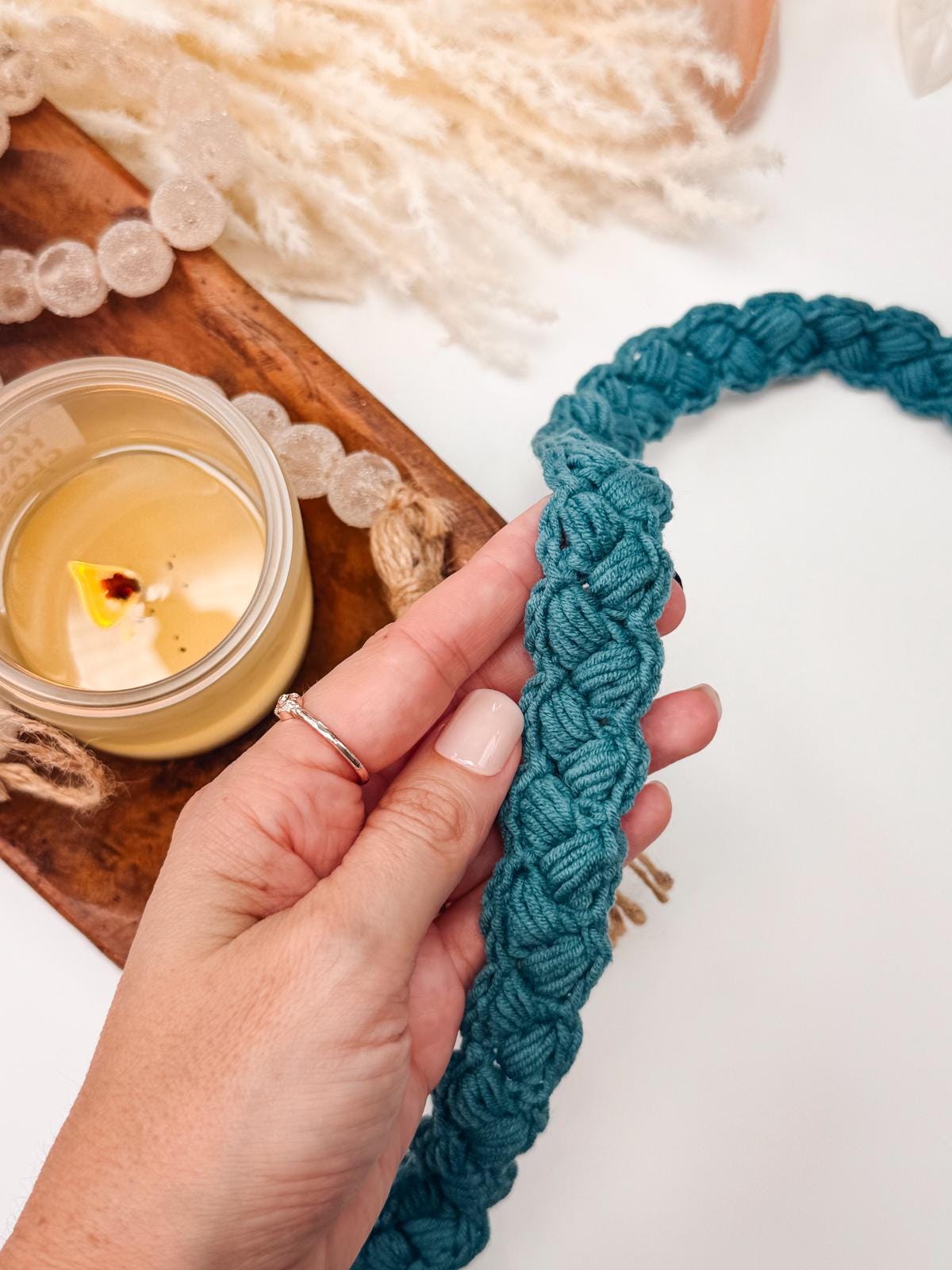 A hand holding a teal braided macramé cord, perfect as a handmade gift, sits next to a lit candle on a wooden tray.