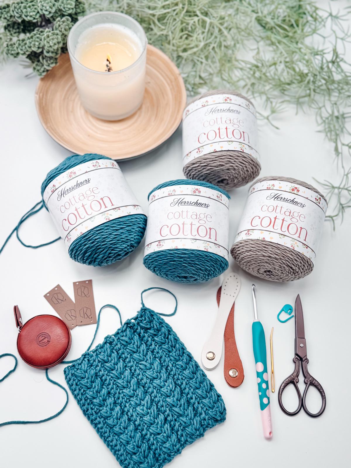Three skeins of yarn, a square swatch, crochet tools, small scissors, a candle, and greenery rest on a white surface—perfect inspiration for creating a handmade gift or pairing with unique jewellery.