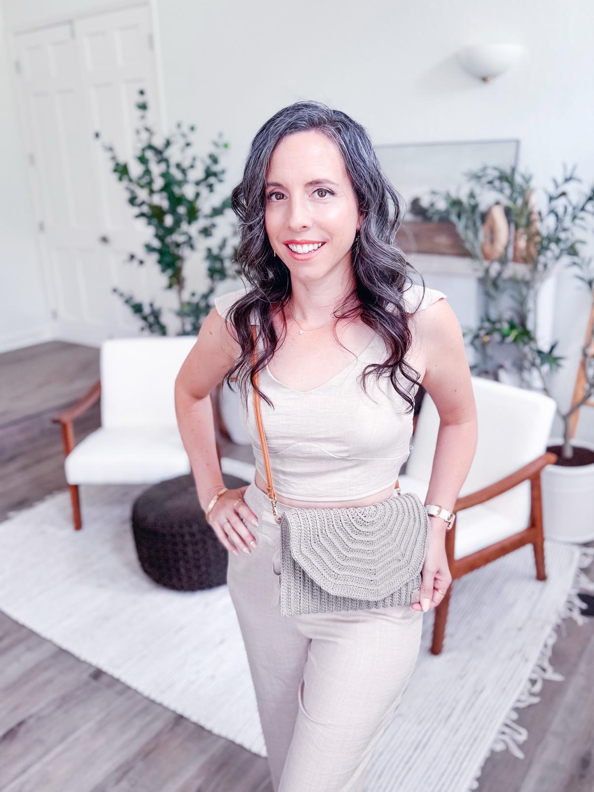 A woman with long wavy hair stands indoors, wearing a light beige sleeveless jumpsuit and holding a crochet crossbody bag, complemented by unique jewellery. White chairs and plants are visible in the background.