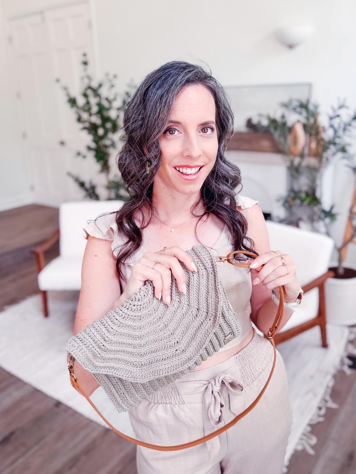 A woman with wavy gray hair stands indoors, holding a woven crochet handbag and smiling at the camera. She wears vintage clothes—a light-colored crop top and high-waisted pants—styled with unique jewellery for a charming, timeless look.