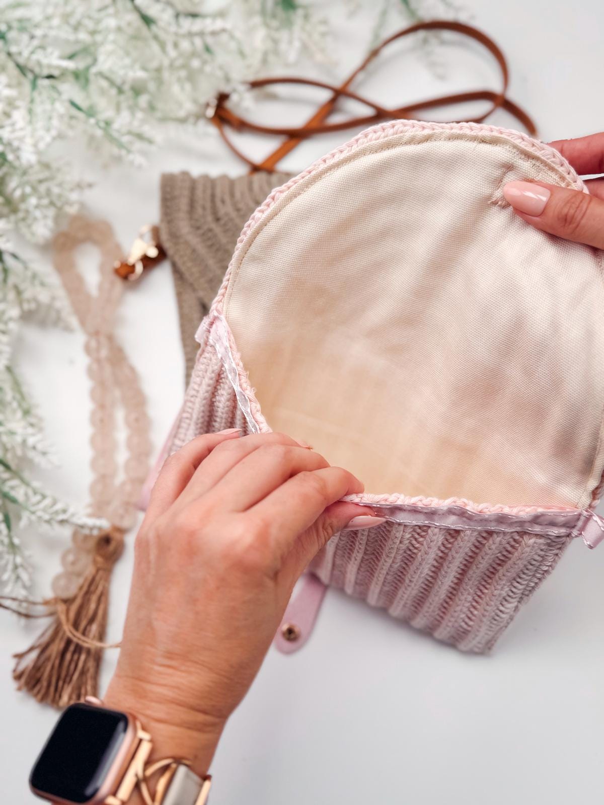 A person opens a small, light pink, crochet handbag with a cream-colored interior lining; unique jewellery and other accessories are visible in the background.