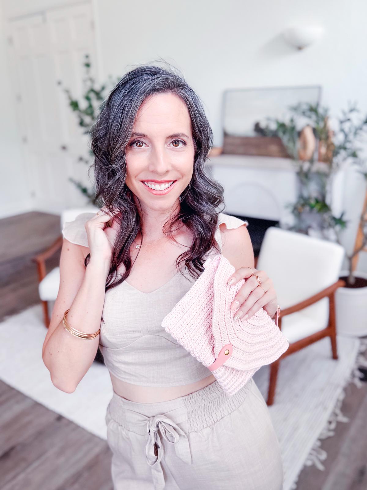 Woman with long wavy hair wearing vintage clothes smiles while holding a pink crochet clutch, standing in a light, modern living room adorned with unique jewellery and handmade gift accents.
