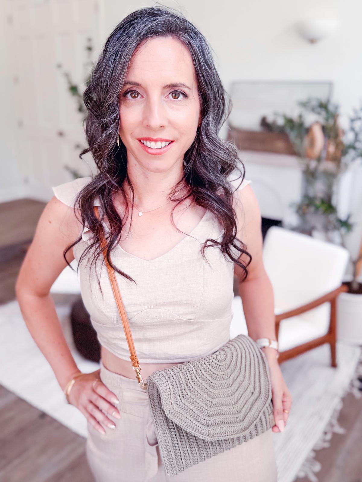 A woman with long wavy dark hair and gray streaks stands indoors, wearing a beige sleeveless outfit, holding a textured beige crochet shoulder bag—the perfect look for shopping vintage clothes or searching for unique jewellery.