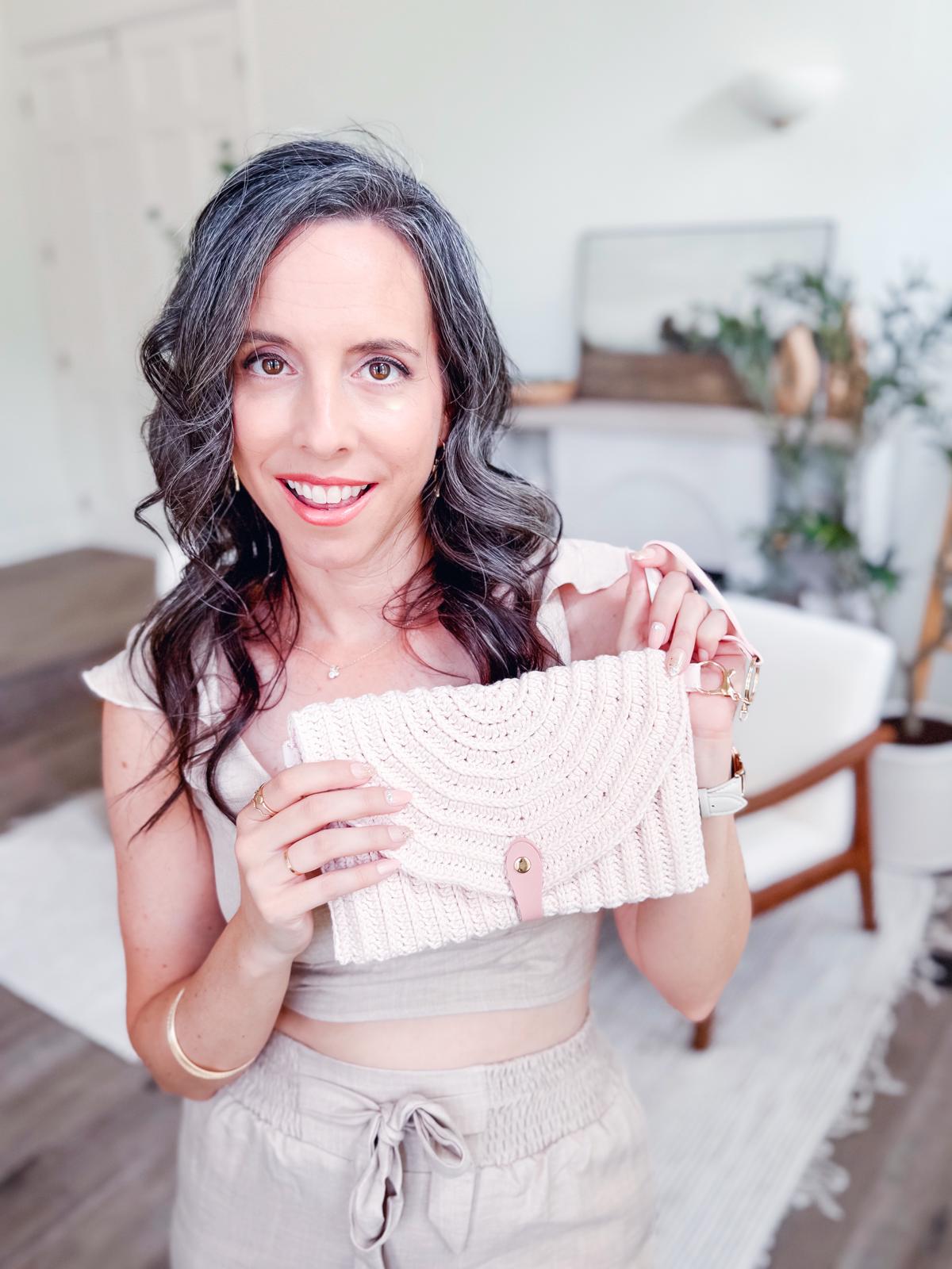 A woman with wavy gray hair stands indoors, holding a woven crochet handbag and smiling at the camera. She wears vintage clothes—a light-colored crop top and high-waisted pants—styled with unique jewellery for a charming, timeless look.