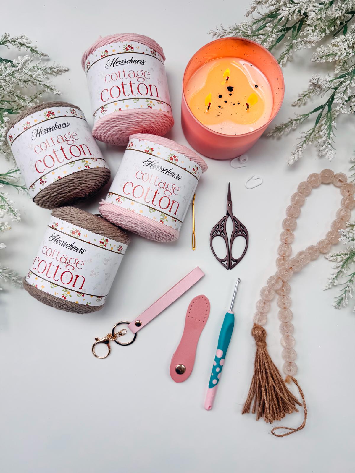 Four skeins of Cottage Cotton yarn, a pink candle, scissors, a beaded tassel, a crochet hook, a keychain, and a yarn cutter arranged on a white surface with greenery—perfect for creating a handmade gift or pairing with unique jewellery.