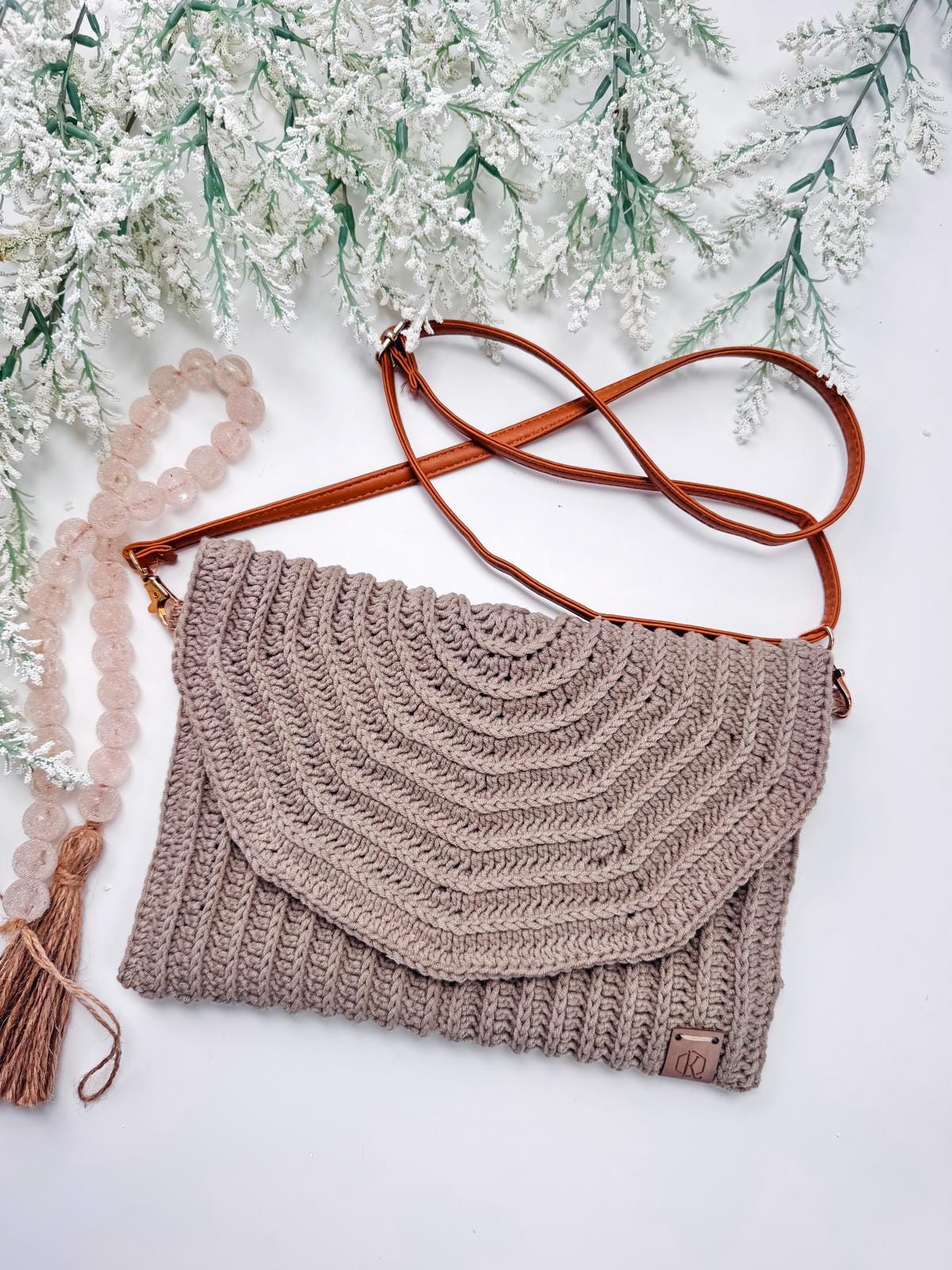 A crocheted brown shoulder bag with a brown strap lies on a white surface beside a beaded tassel accessory and frosted foliage—perfect as a handmade gift or to complement your unique jewellery.