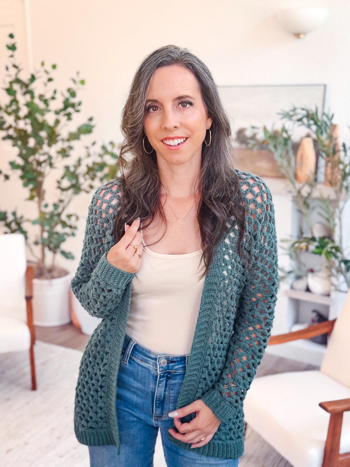 Woman with long brown hair wearing a green open-crochet cardigan, white top, unique jewellery, and jeans stands indoors in a room with plants and white chairs.