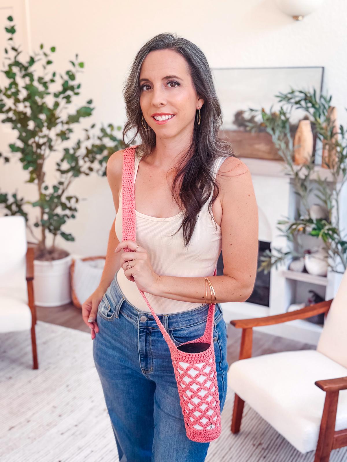 A woman in a beige tank top and jeans stands indoors, holding a pink crocheted bottle holder over her shoulder. She is smiling, surrounded by plants and chairs—her look accented with unique jewellery, making it a perfect handmade gift moment.