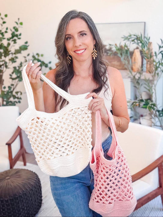 A woman stands indoors holding two crocheted tote bags, one white and one pink—perfect as a handmade gift—amidst plants and chairs in the background.