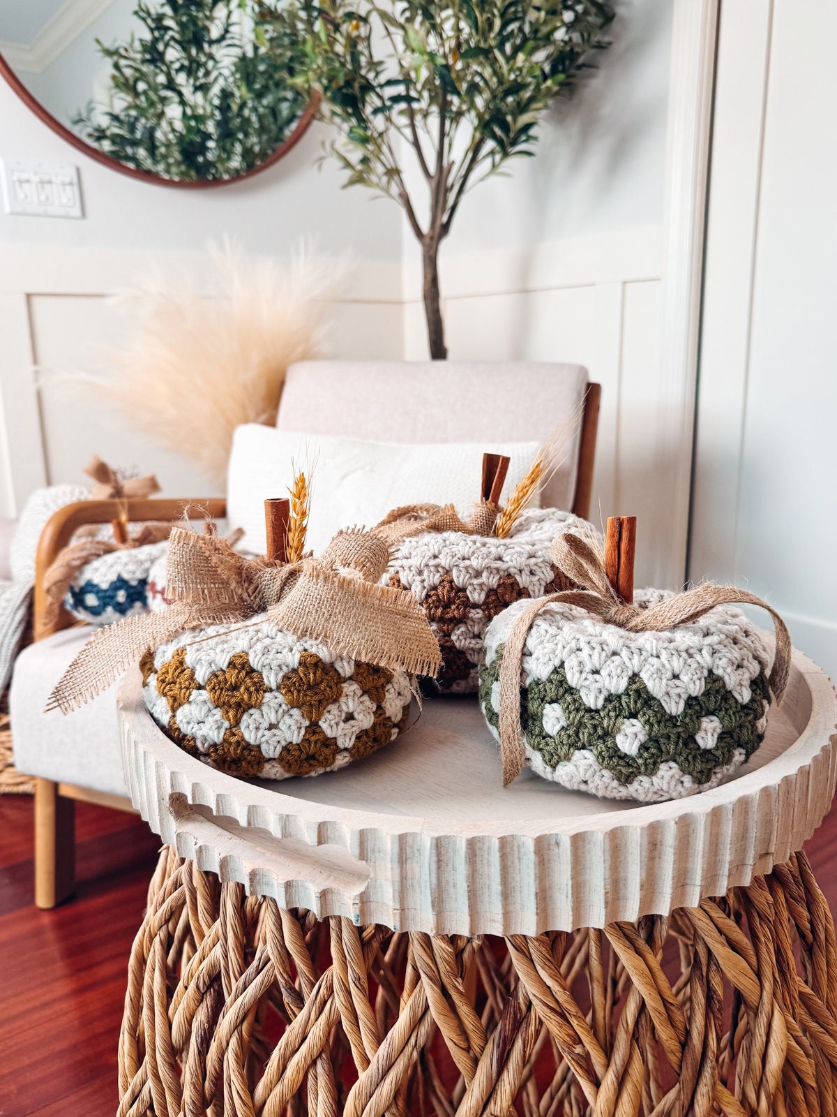 Three crocheted pumpkins with burlap bows and cinnamon stick stems sit on a round wicker table. A beige chair, potted plant, and vintage clothes create a charming background scene.