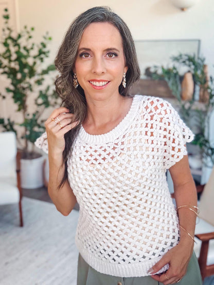 A woman with long brown hair wearing a white crocheted top and unique jewellery stands indoors, smiling at the camera. There are plants and chairs in the background.
