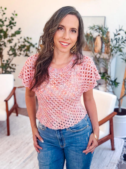 A woman with long brown hair wears a pink crochet top and blue jeans, standing indoors with plants, white chairs, and unique jewellery in the background.
