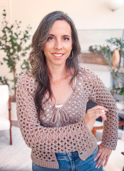A woman with long brown hair and a crochet sweater smiles at the camera indoors, standing in a bright room with plants, neutral decor, and unique jewellery accenting her style.
