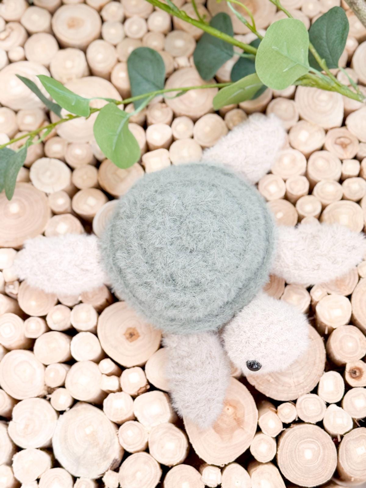 A plush turtle toy with a green shell and gray limbs lies on a surface covered with round wooden logs and green leaves, making a charming handmade gift.