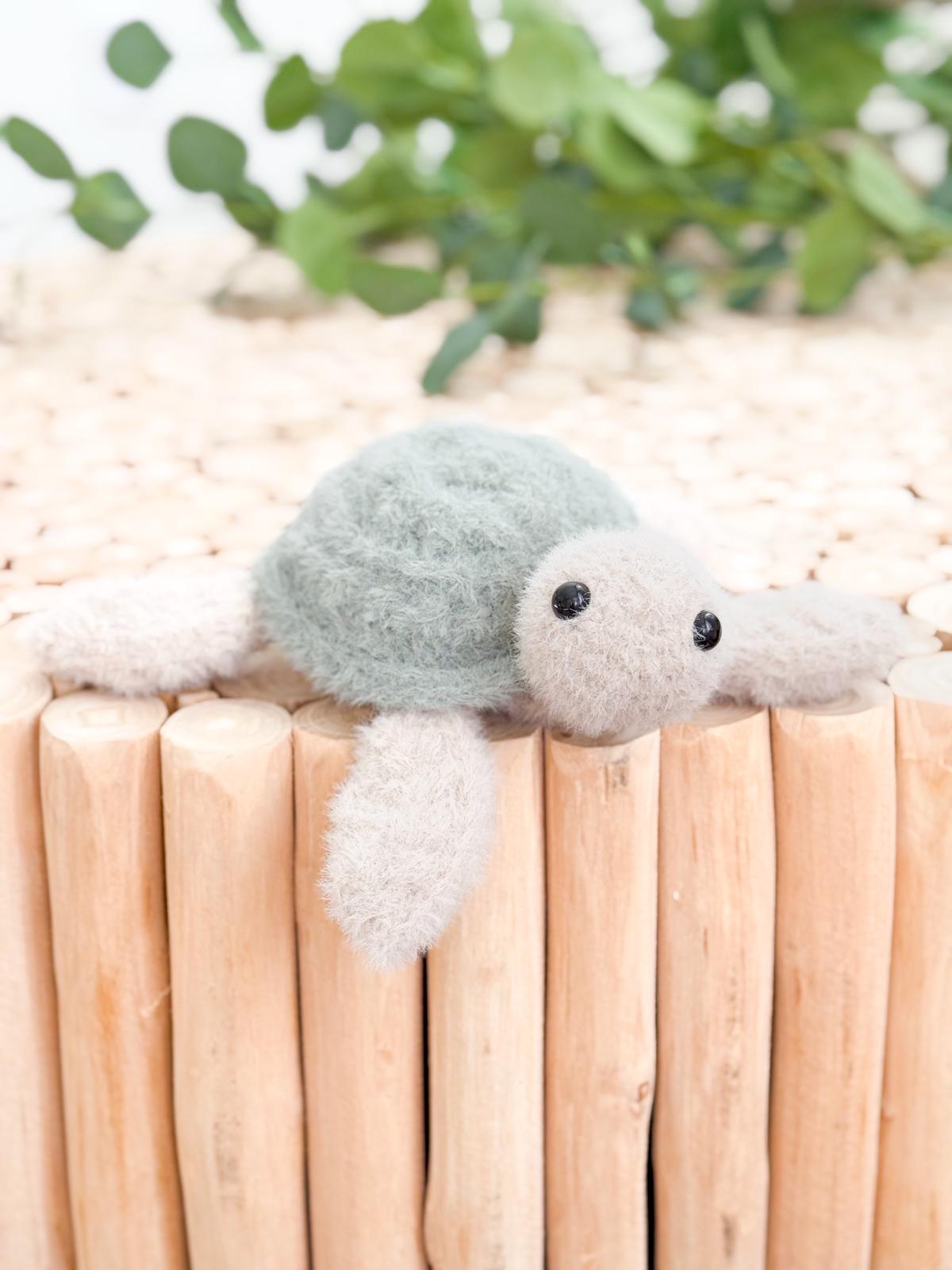 A small, plush turtle toy with a green shell and beige limbs rests on a wooden surface, making a charming handmade gift, with green foliage in the background.