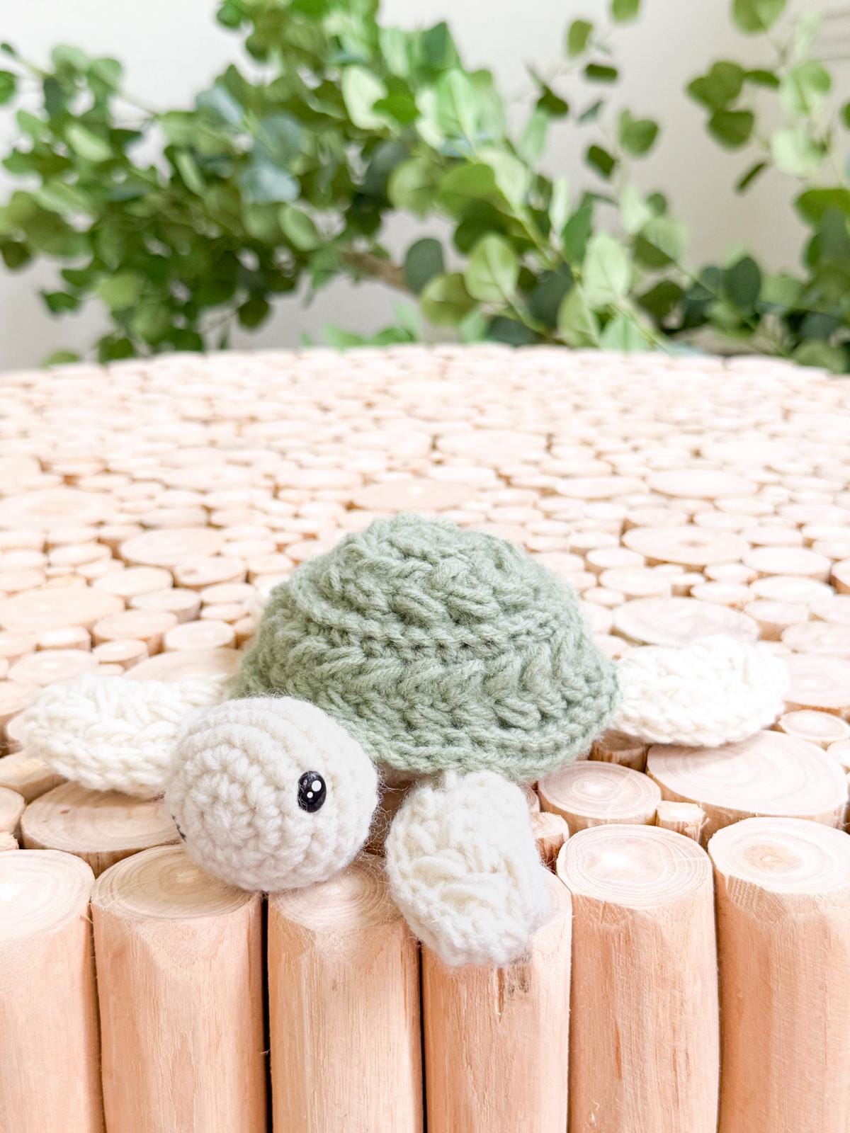 A crocheted turtle with a green shell and white limbs rests on stacked wooden sticks, leafy plants in the background—a charming handmade gift perfect for nature lovers.