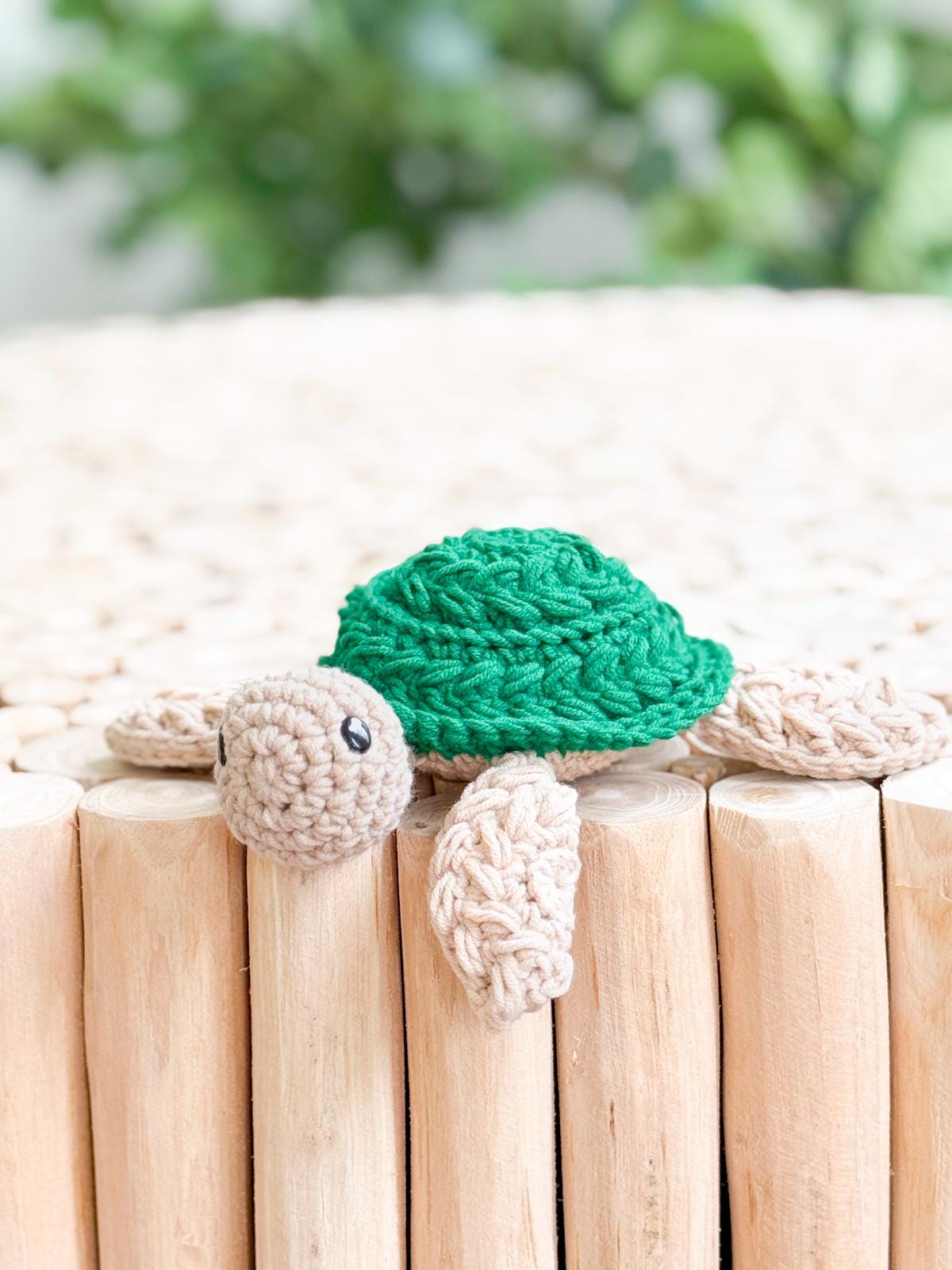 A crocheted turtle with a green shell and beige limbs rests on a surface made of vertical wooden logs, making a delightful handmade gift, with greenery blurred in the background.
