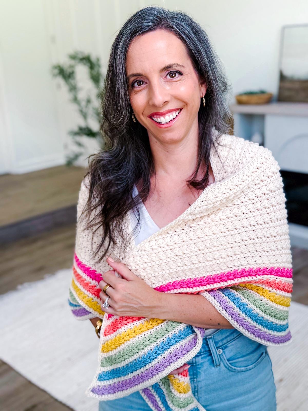 A woman with long brown hair and gray streaks smiles while wearing a cream-colored crochet shawl with rainbow stripes on the edge, standing in a bright, modern room—her look completed by unique jewellery.