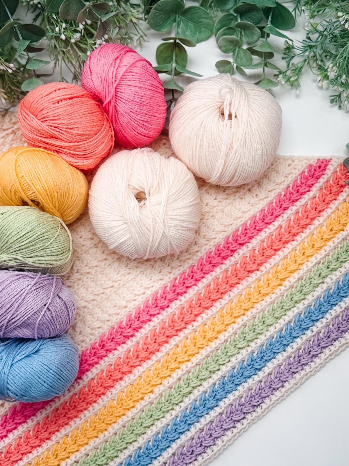 Six balls of colorful yarn are arranged next to a partially completed crochet project with striped pastel rows, set against green leaves—perfect inspiration for a handmade gift.