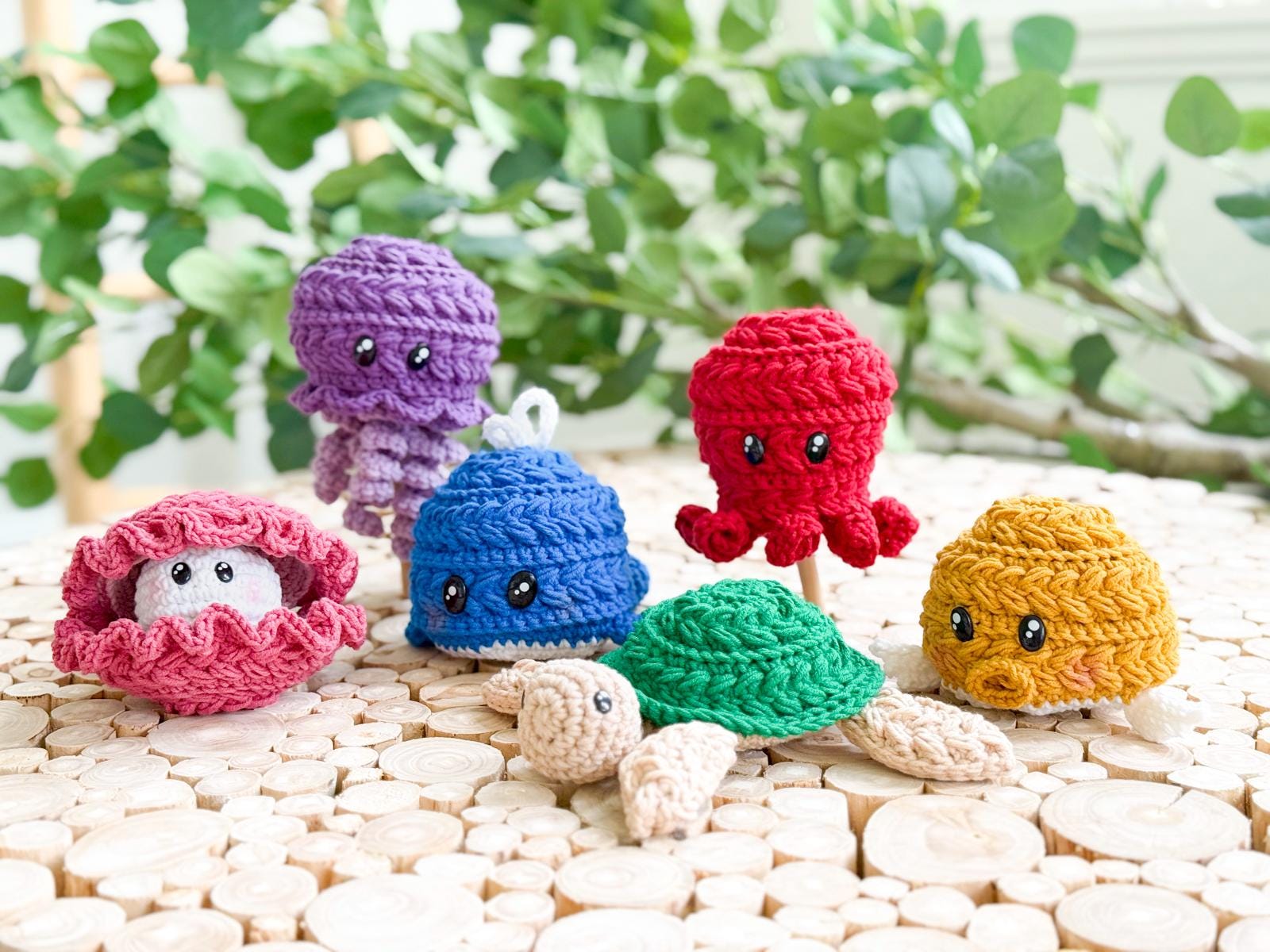 Five colorful crocheted sea creature toys, including a clam, jellyfish, octopus, and turtle, are displayed on a wooden surface with green plants in the background—an adorable handmade gift for any ocean lover.