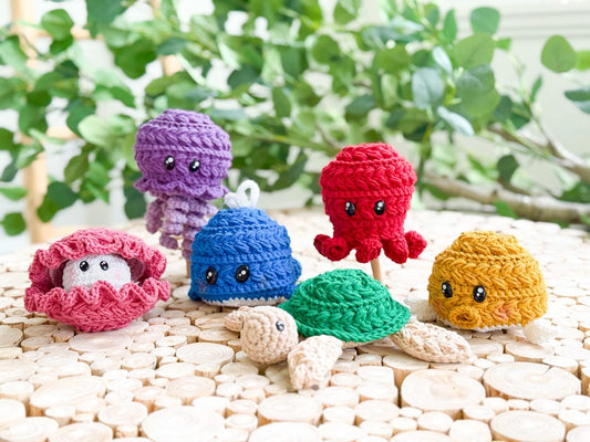 Five colorful crocheted sea creature toys, including a clam, jellyfish, octopus, and turtle, are displayed on a wooden surface with green plants in the background—an adorable handmade gift for any ocean lover.