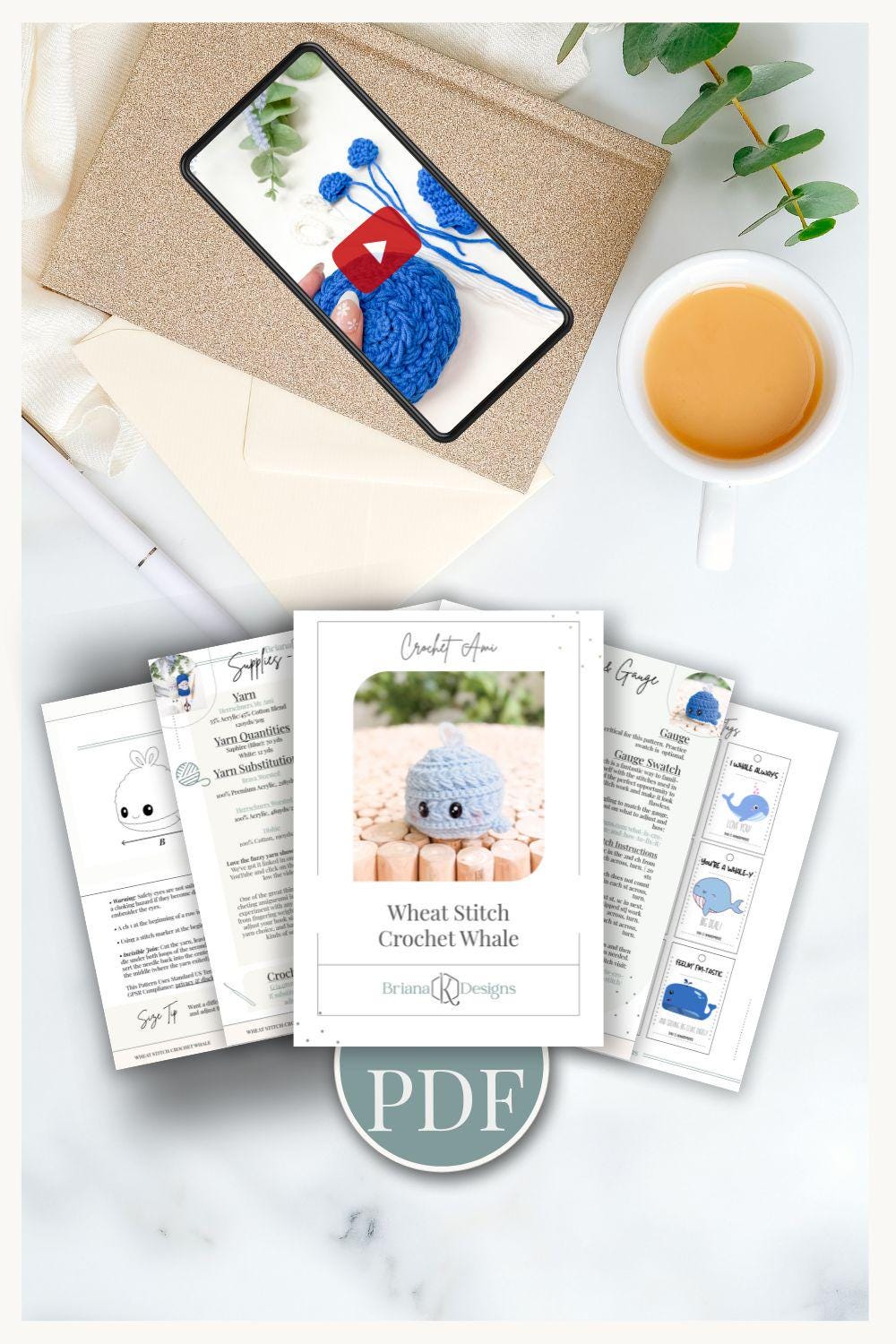A cup of tea, an envelope with a phone showing blue yarn, unique jewellery, and printed crochet whale pattern pages labeled "PDF" are arranged on a marble surface.