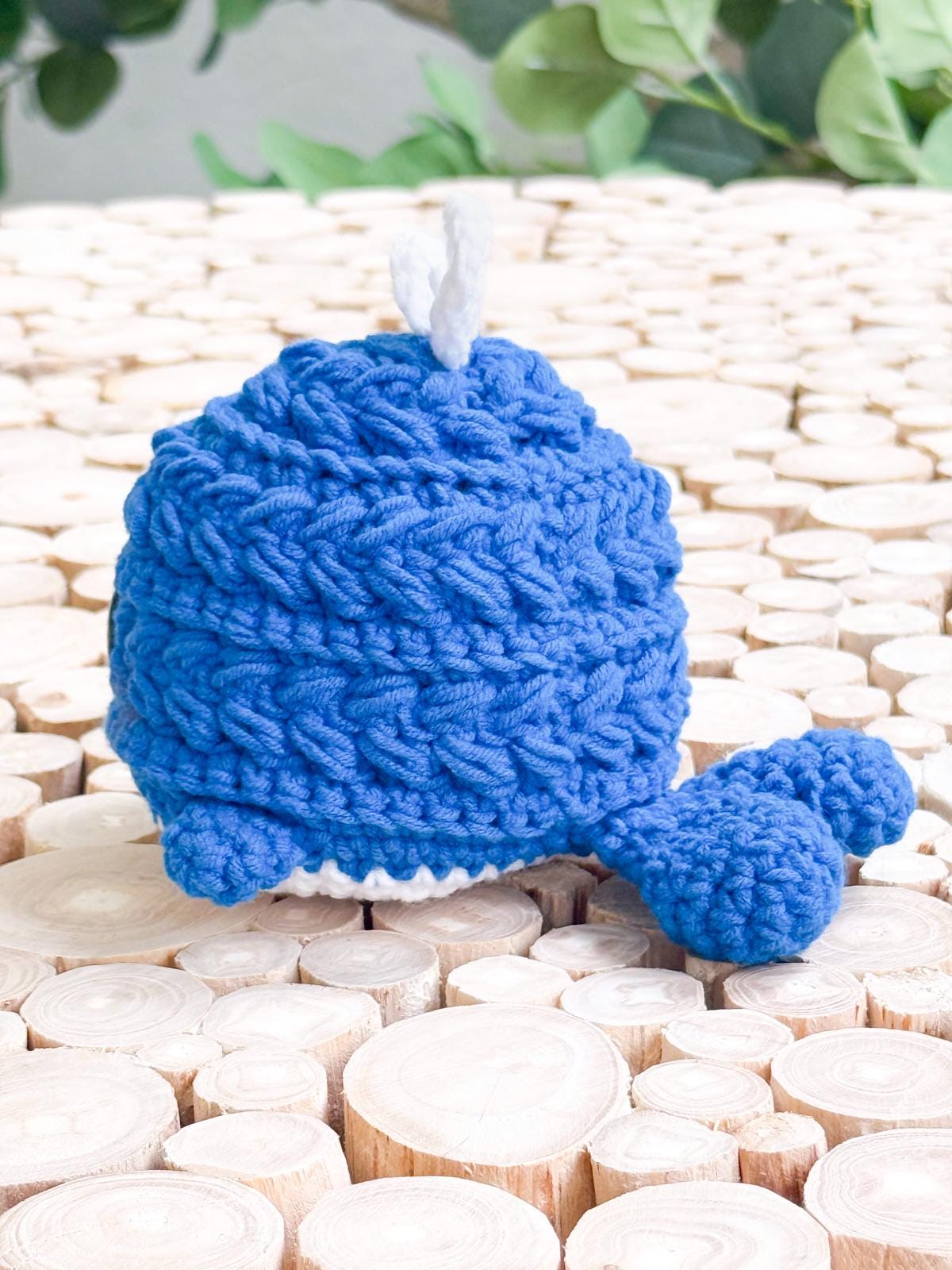 A blue crocheted whale with a white spout sits on a surface made of cut wooden logs, with green leaves in the background—perfect as a charming handmade gift.