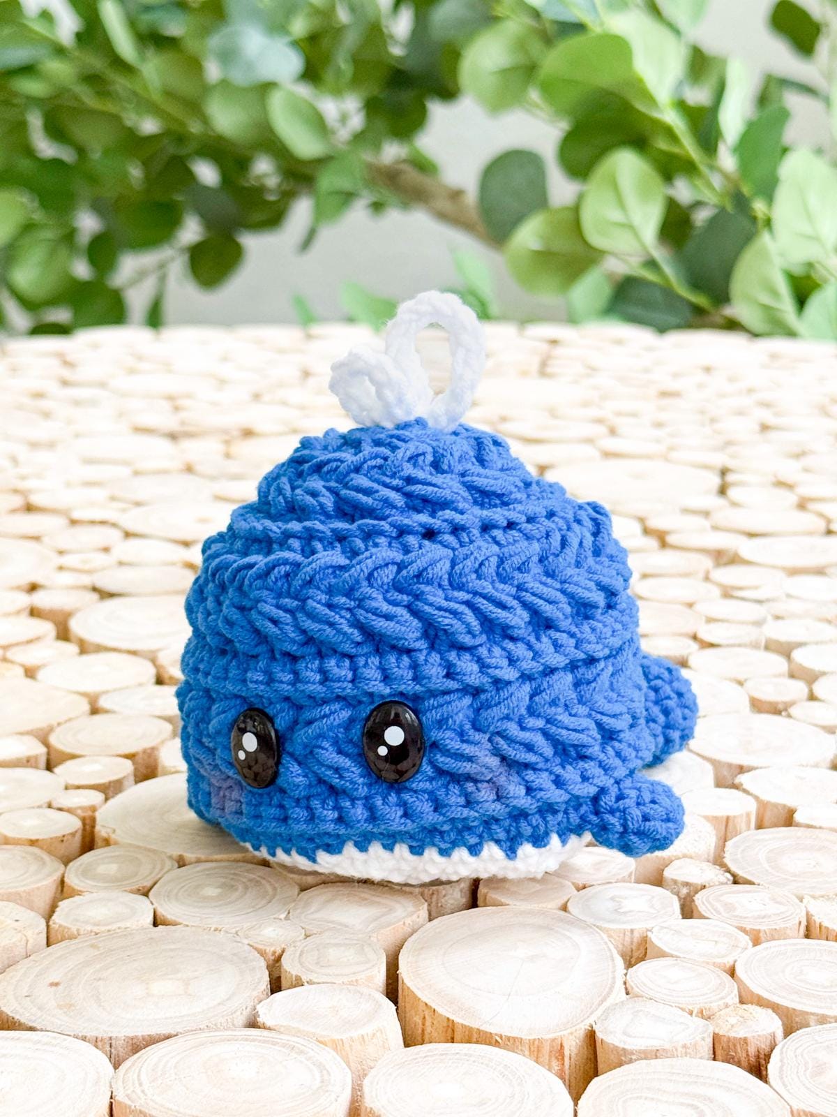 A small, blue crocheted whale with black button eyes sits on a surface of light wooden discs, making a charming handmade gift. Green leafy plants in the background add to its appeal.
