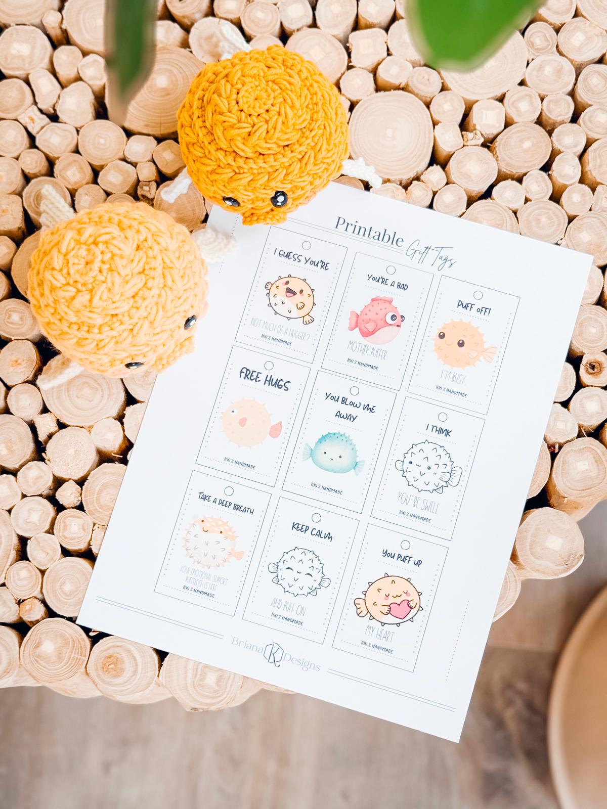 A sheet of printable gift tags with illustrated animals and puns lies on a wooden circular-pattern table beside two yellow crocheted amigurumi fish.