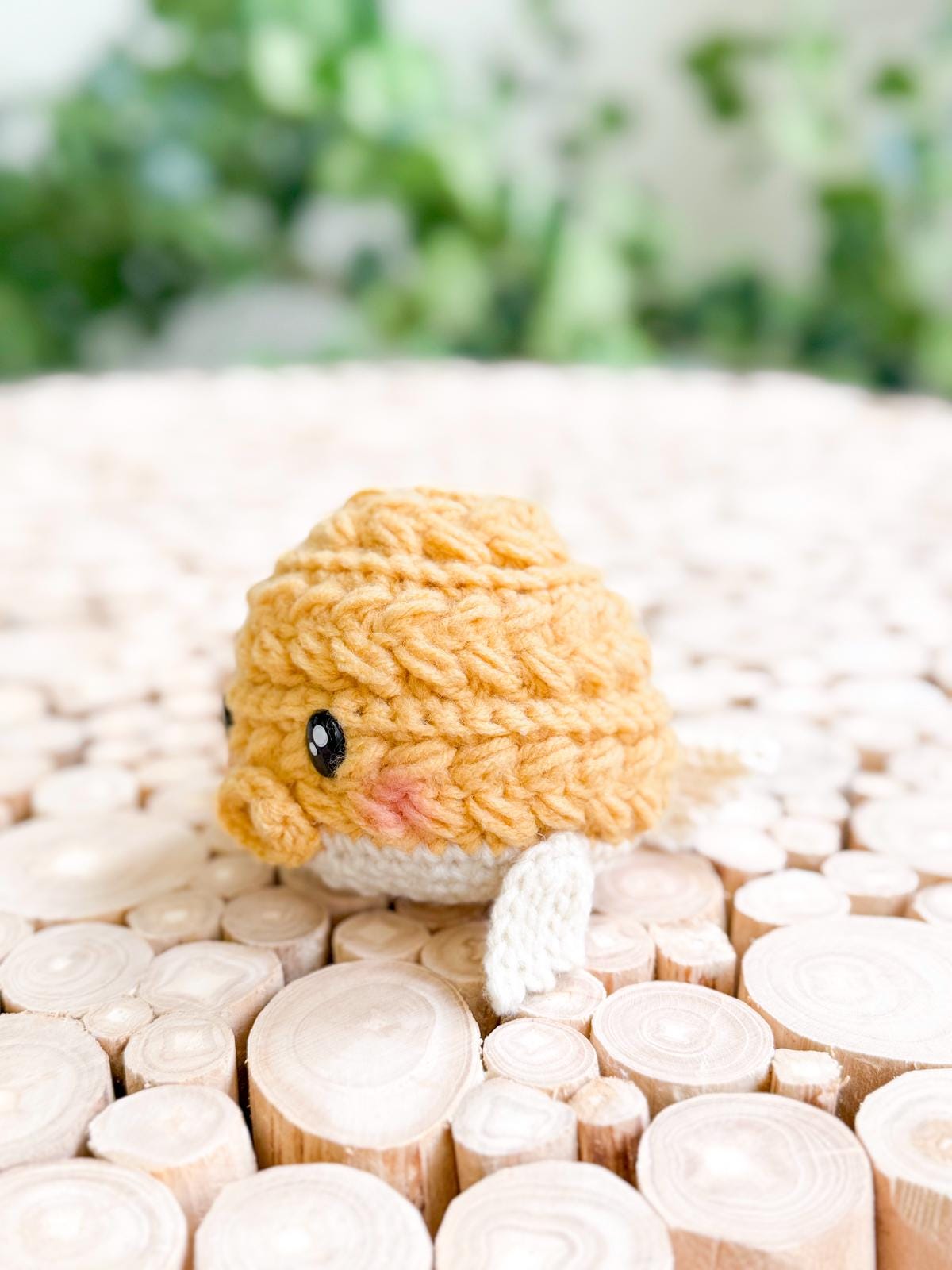 A small, yellow, crocheted fish plush with white fins sits on a surface made of circular wood slices, with green foliage blurred in the background—an adorable handmade gift perfect for any occasion.