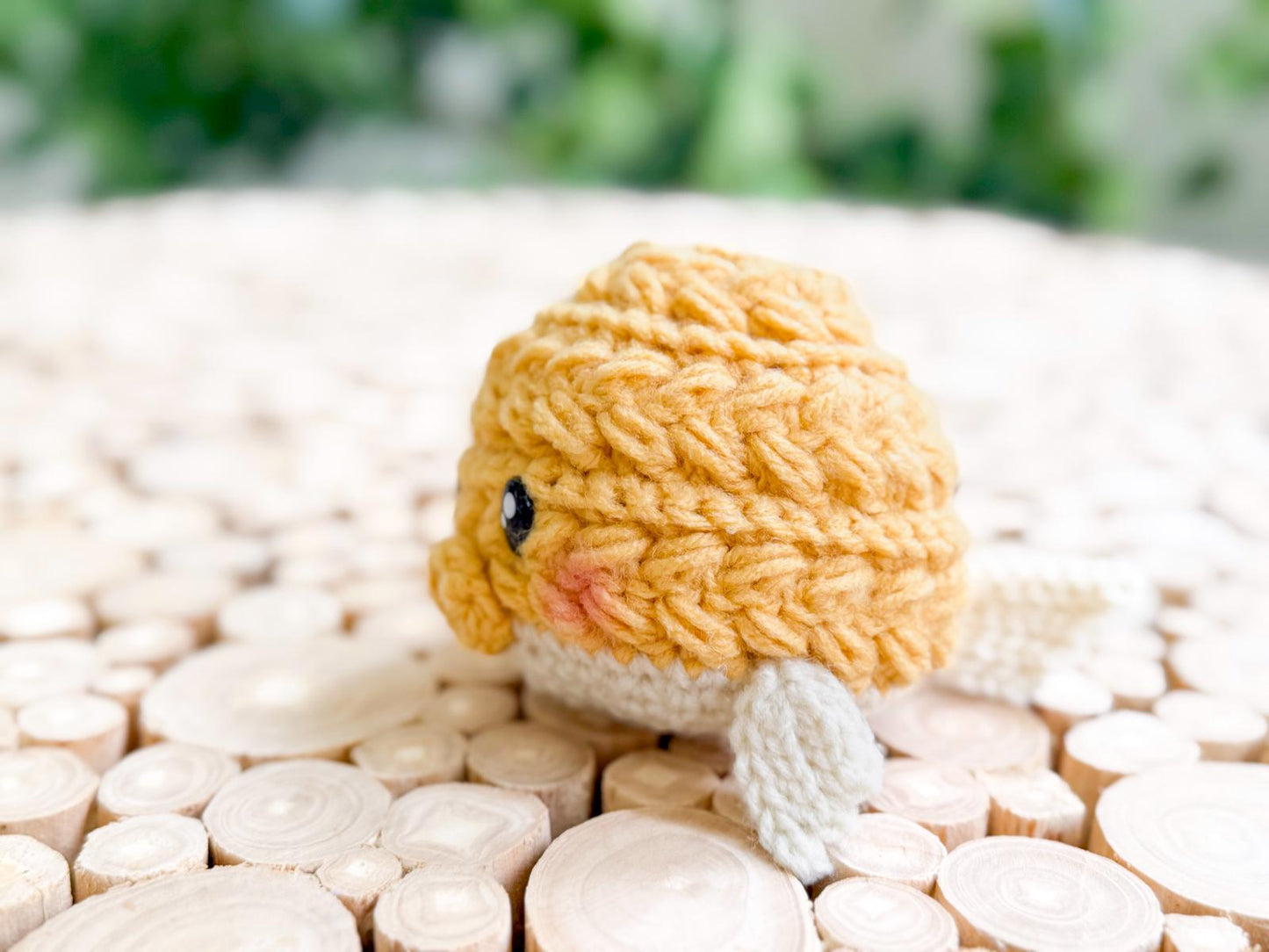 A small, crocheted yellow and white fish with black eyes and pink cheeks rests on a surface made of circular wooden pieces—perfect as a charming handmade gift.