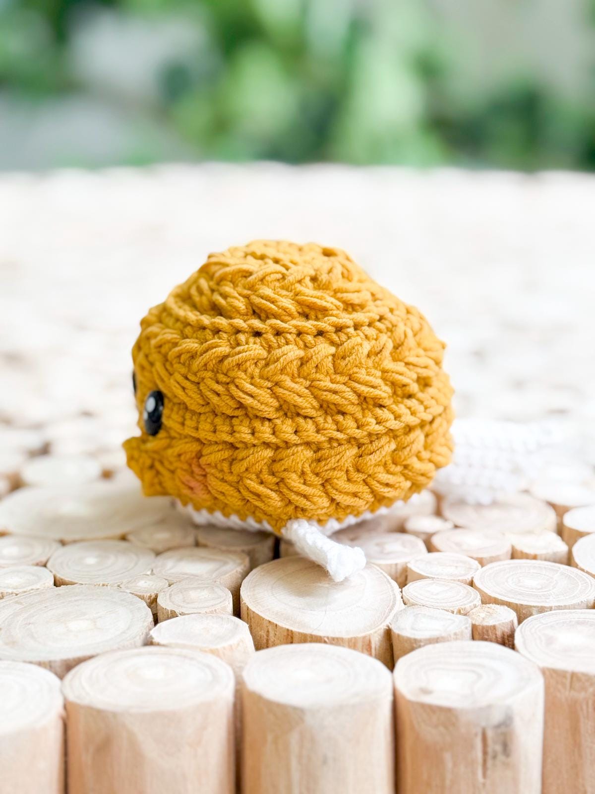 A small, round, yellow crocheted toy with white appendages sits on a surface made of cut wooden logs. Crochet amigurumi pufferfish.