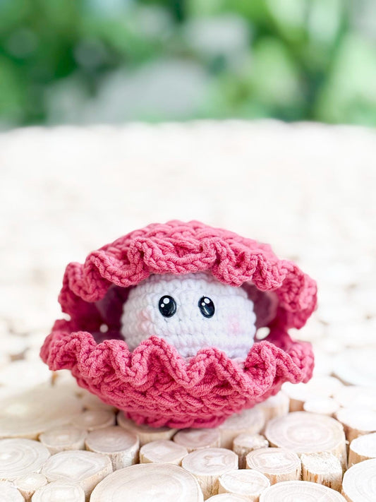 A crocheted pink clam with a smiling face peeks out from its shell, set on a light wooden surface with a blurred green background—a charming handmade gift perfect for fans of unique jewellery and whimsical decor.
