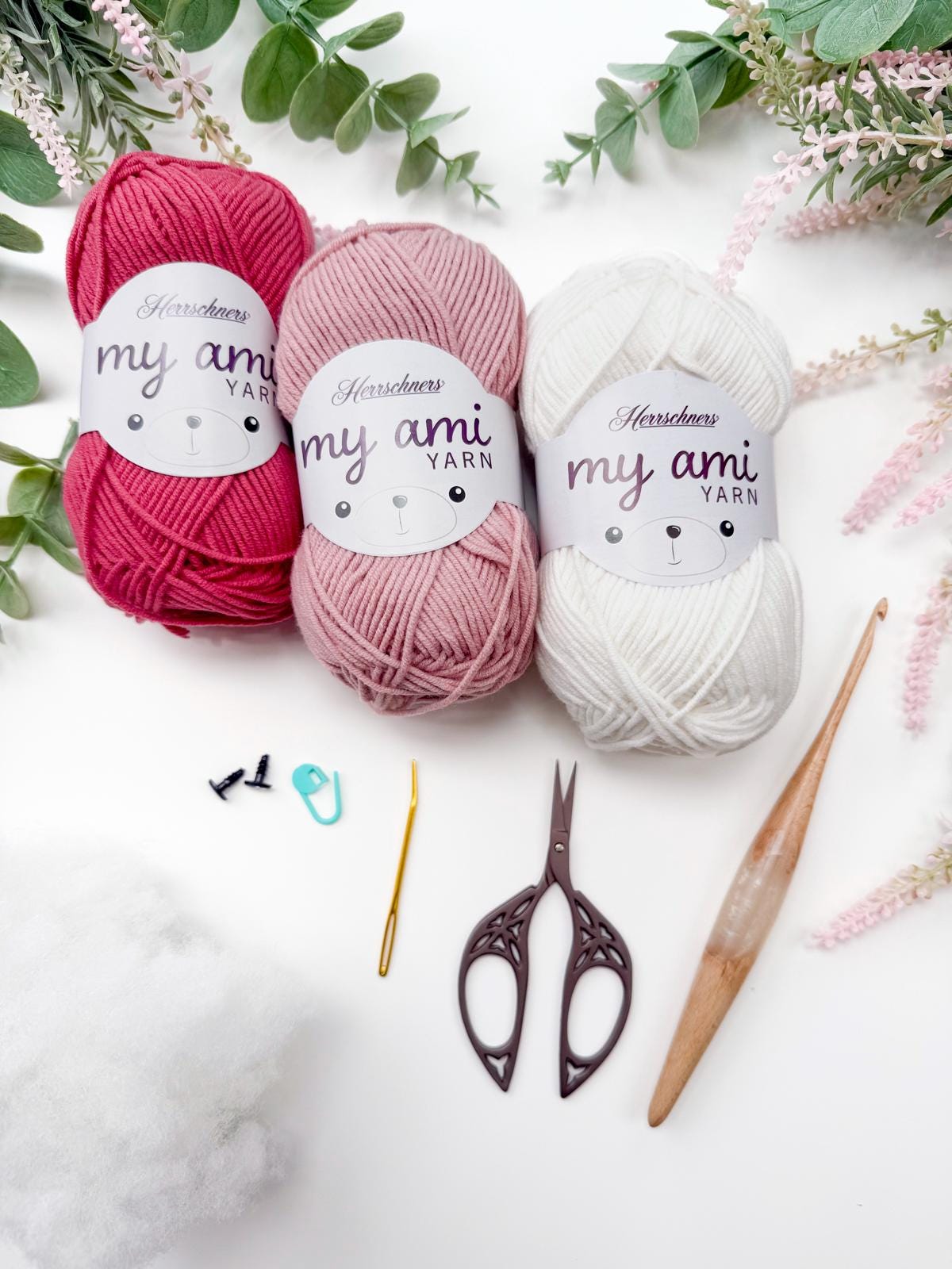 Three skeins of yarn in red, pink, and white with crochet tools, safety eyes, a stitch marker, and stuffing—perfect for crafting a handmade gift—arranged on a white surface with greenery.