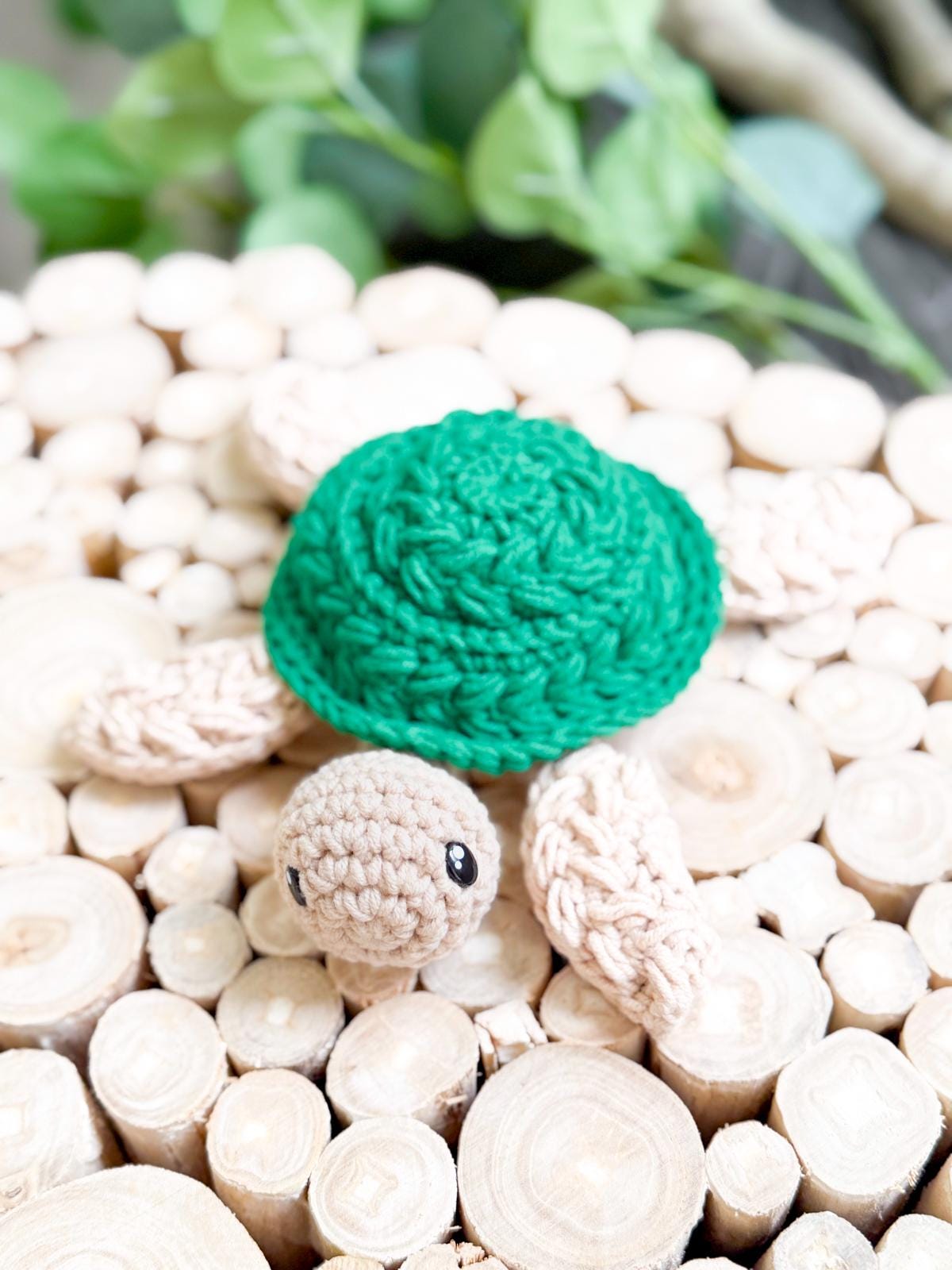 A crocheted turtle toy with a green shell and beige limbs is displayed on a surface made of cut wooden logs, making it a charming handmade gift.