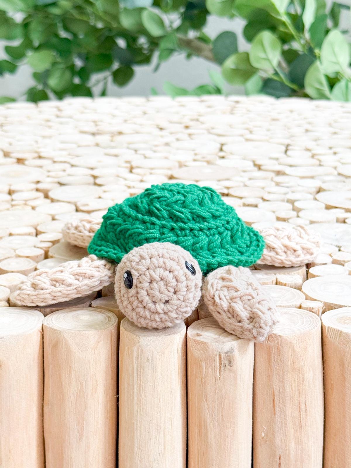 A crocheted turtle with a green shell and beige limbs is placed on a surface made of vertical wooden dowels, with leafy greenery in the background—a charming handmade gift for any occasion.