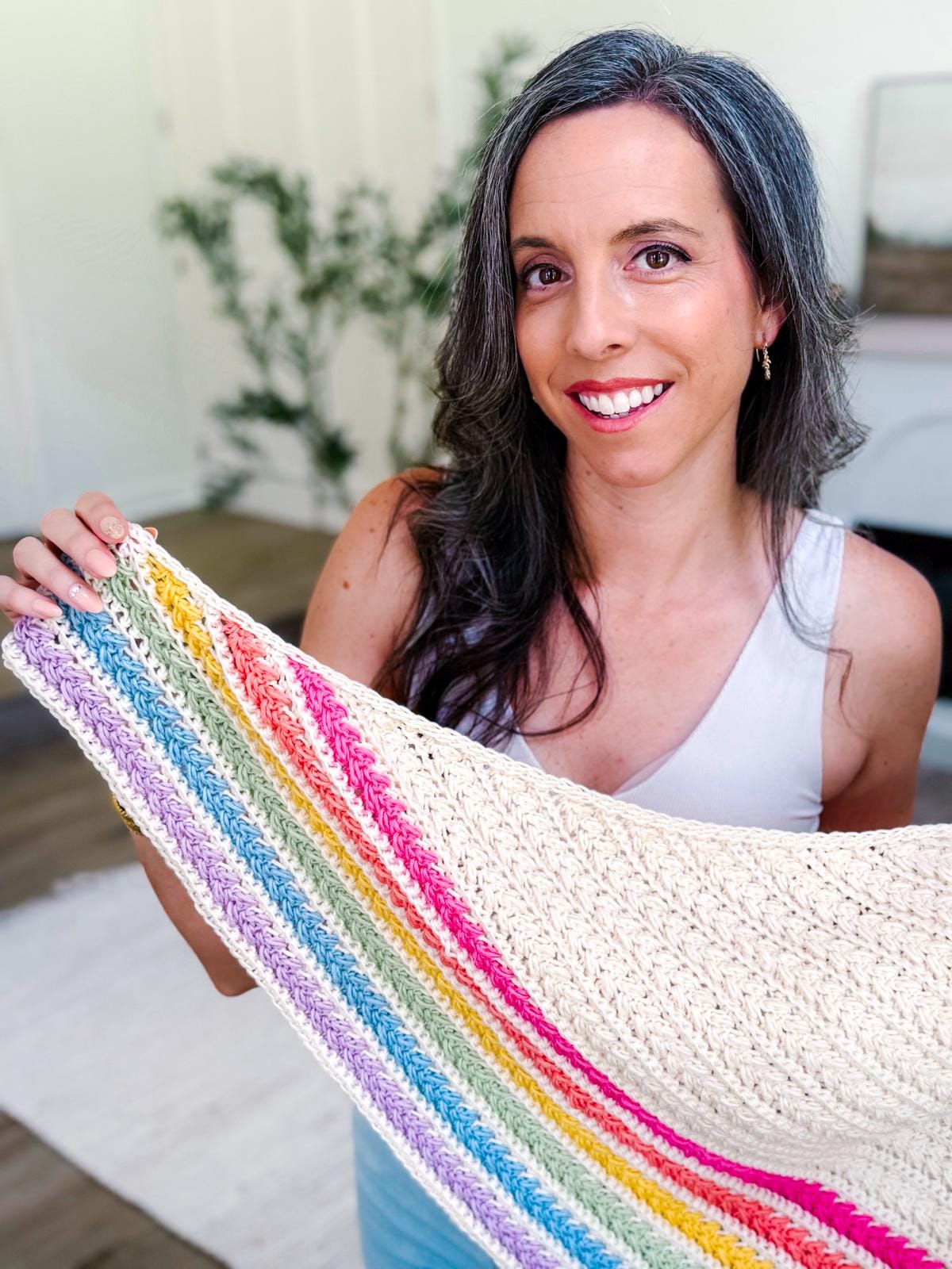A woman with long brown hair wearing a colorful crocheted shawl and unique jewellery stands in a bright room with a white rug and potted plant.